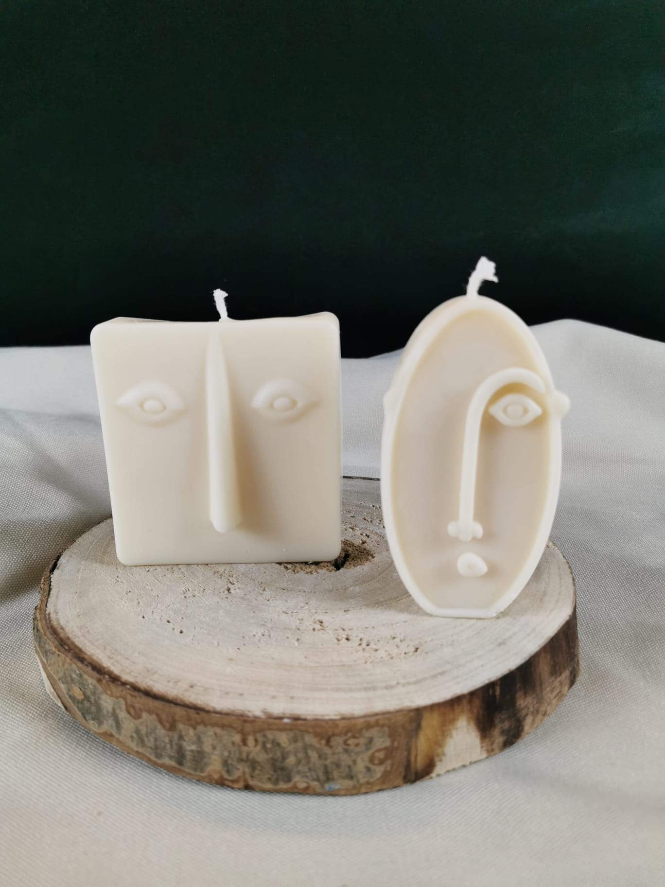 Zwei handgegossene abstrakte Gesichts-Kerzen aus hellem Rapswachs, eine quadratisch und eine oval, dekorativ auf einer Holzscheibe platziert.