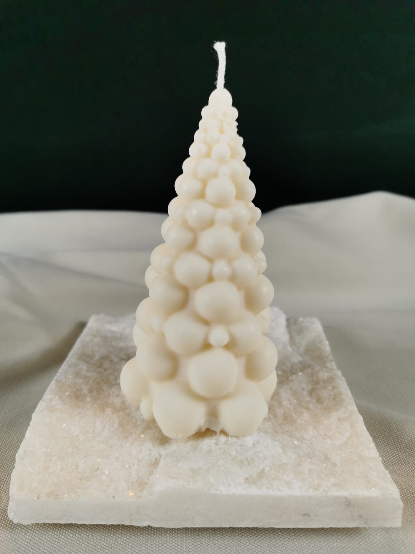 Handgegossene Weihnachtsbaumkerze aus hellem Rapswachs mit kugelförmiger Struktur, dekorativ auf einer glitzernden Steinplatte präsentiert.