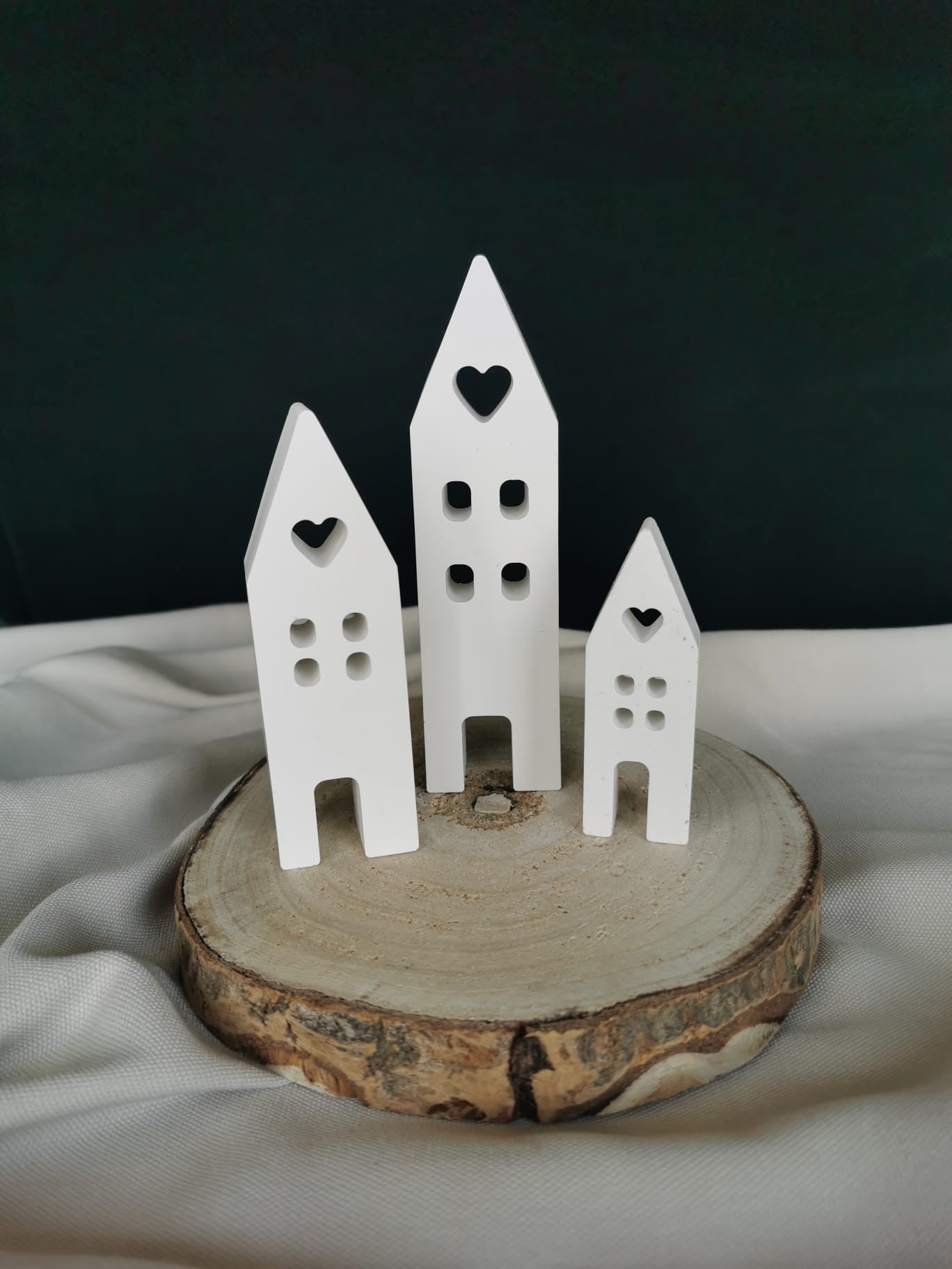 Weiße Deko-Häuser aus Gießkeramik im 3er Set, mit ausgestanzten Herzfenstern und minimalistischer Form, dekorativ auf einer Holzscheibe platziert.