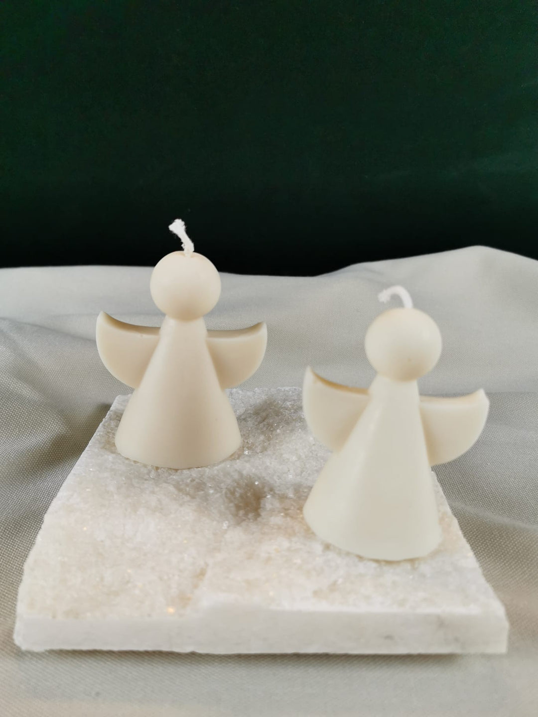 Zwei handgegossene Engel-Figurenkerzen aus hellem Rapswachs, mit runden Köpfen und ausgebreiteten Flügeln, dekorativ auf einer glitzernden Steinplatte präsentiert.