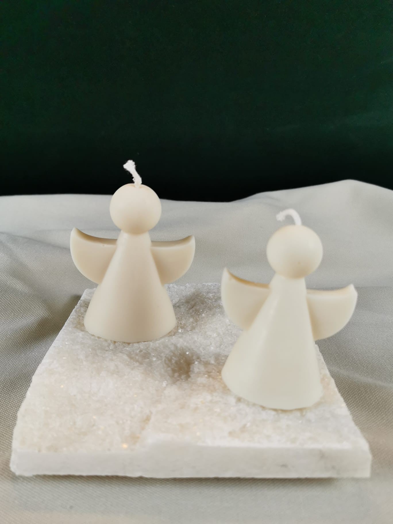 Zwei handgegossene Engel-Figurenkerzen aus hellem Rapswachs, mit runden Köpfen und ausgebreiteten Flügeln, dekorativ auf einer glitzernden Steinplatte präsentiert.