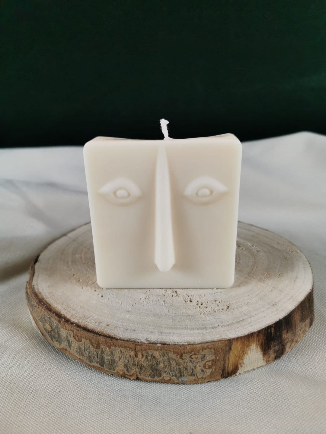 Handgegossene abstrakte Gesichts-Kerze aus hellem Rapswachs in quadratischer Form, auf einer Holzscheibe dekorativ präsentiert.