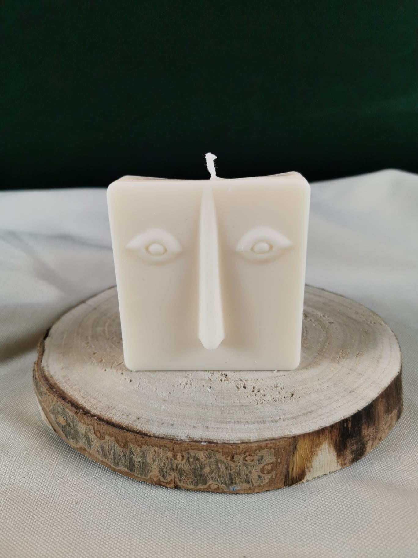 Handgegossene abstrakte Gesichts-Kerze aus hellem Rapswachs in quadratischer Form, auf einer Holzscheibe dekorativ präsentiert.