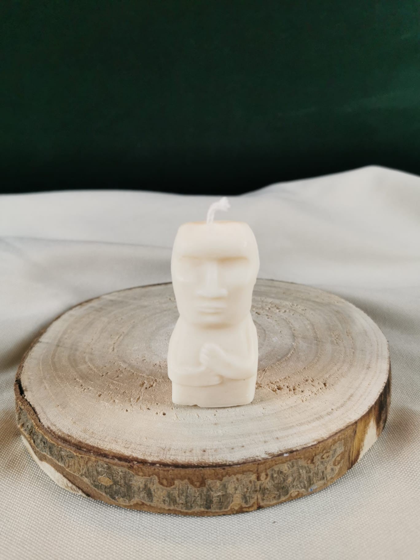 Handgegossene Figurenkerze aus hellem Rapswachs, die wie eine stilisierte sitzende Figur mit gefalteten Händen gestaltet ist. Präsentiert auf einer Holzscheibe.