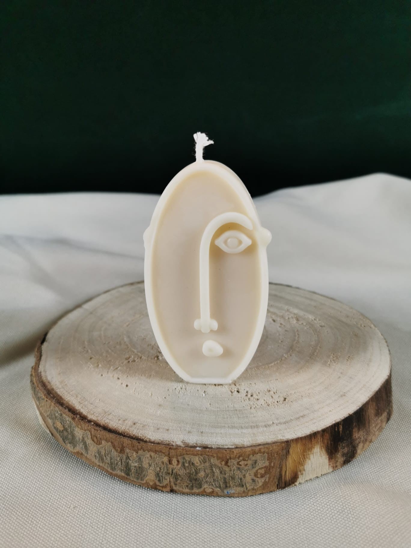 Handgegossene ovale Figurenkerze aus hellem Rapswachs mit abstrakter Gesichtslinie, dekorativ auf einer Holzscheibe präsentiert.