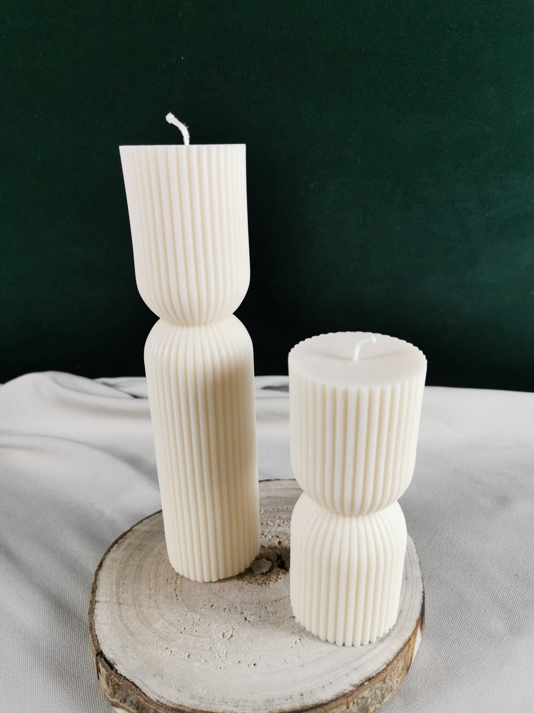 „Zwei handgegossene, cremefarbene Rapswachskerzen im modernen geriffelten Design, fotografiert auf einer Holzscheibe – große und kleine Version.“