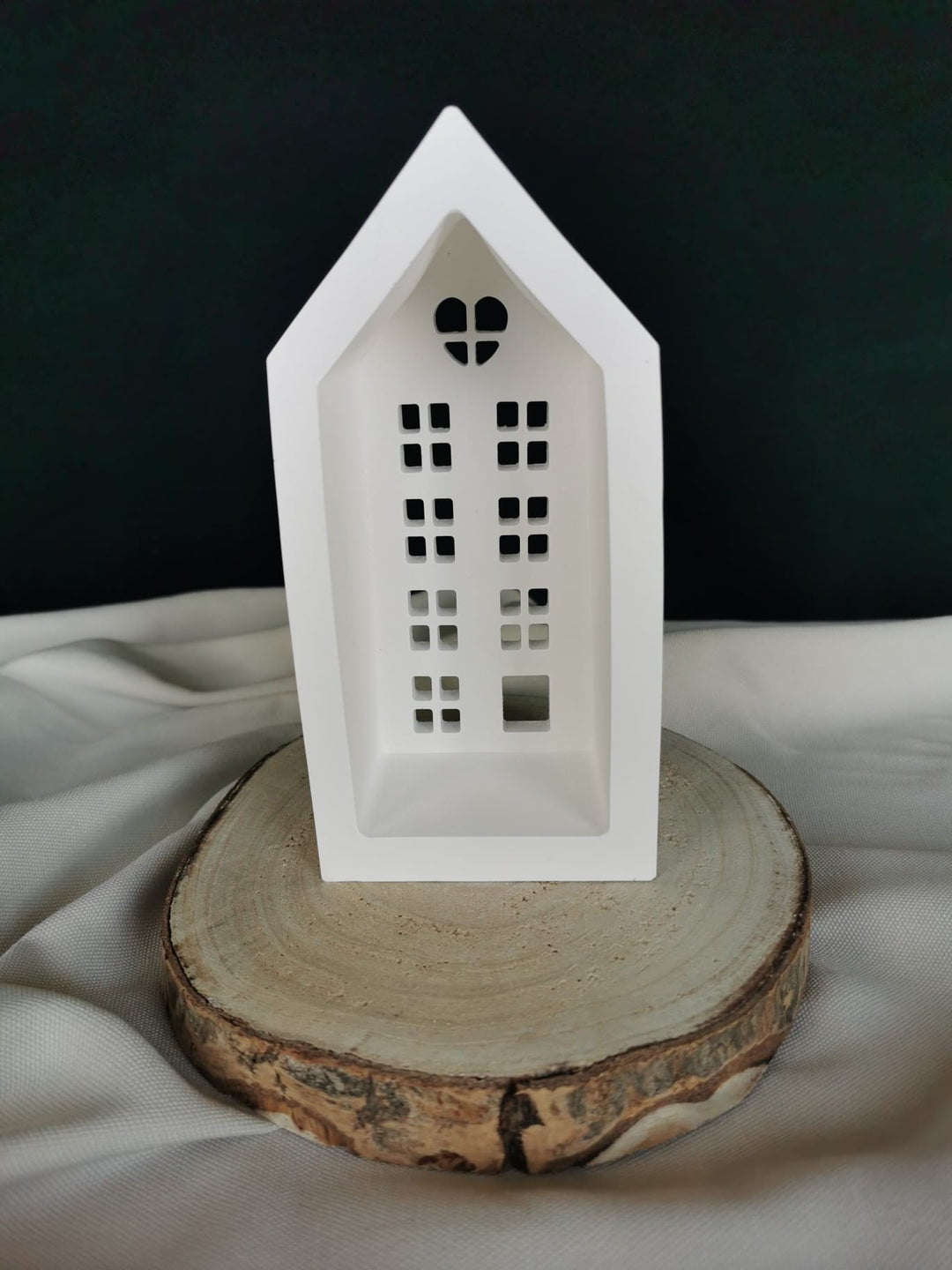 Weiße Keramik-Teelichthaus-Dekoration mit ausgestanzten Fenstern und Herzfenster, auf einer Holzscheibe präsentiert – winterliche Weihnachtsdeko.