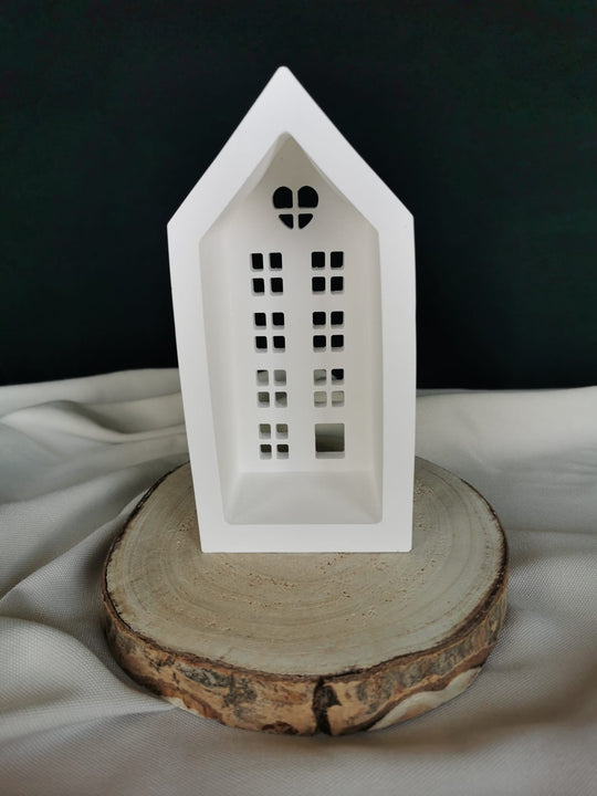 Weiße Keramik-Teelichthaus-Dekoration mit ausgestanzten Fenstern und Herzfenster, auf einer Holzscheibe präsentiert – winterliche Weihnachtsdeko.