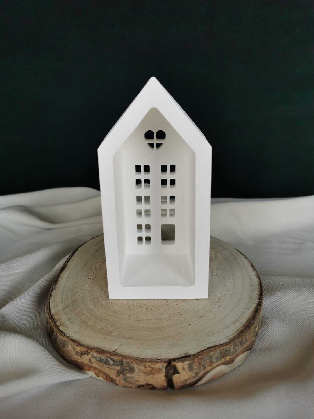 Weiße Keramik-Teelichthaus-Dekoration mit ausgestanzten Fenstern und Herzfenster, auf einer Holzscheibe präsentiert – winterliche Weihnachtsdeko.