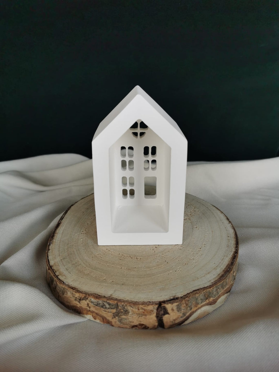 Weiße Keramik-Teelichthaus-Dekoration mit ausgestanzten Fenstern und Herzfenster, auf einer Holzscheibe präsentiert – winterliche Weihnachtsdeko.