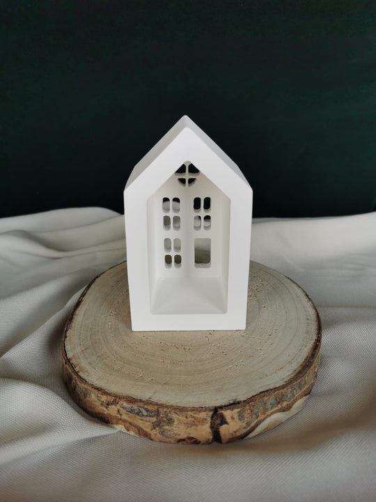 Weiße Keramik-Teelichthaus-Dekoration mit ausgestanzten Fenstern und Herzfenster, auf einer Holzscheibe präsentiert – winterliche Weihnachtsdeko.