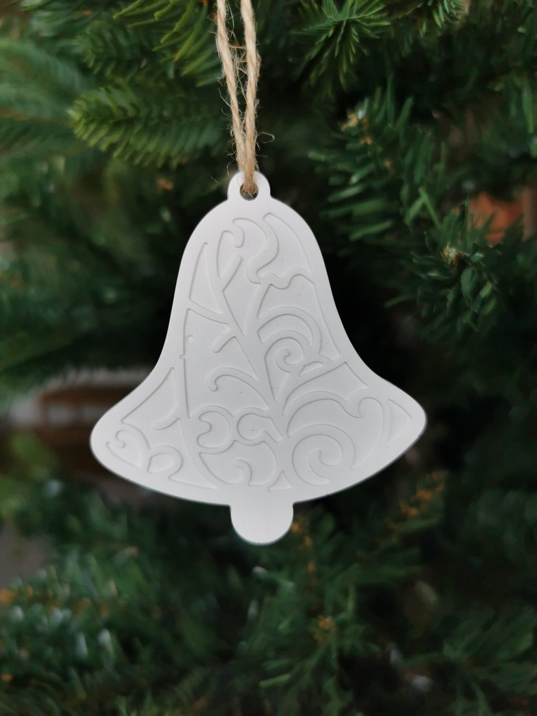 „Weißer Keramik-Baumanhänger in Glockenform mit Ornamentmuster, aufgehängt an einer Juteschnur im Weihnachtsbaum.“