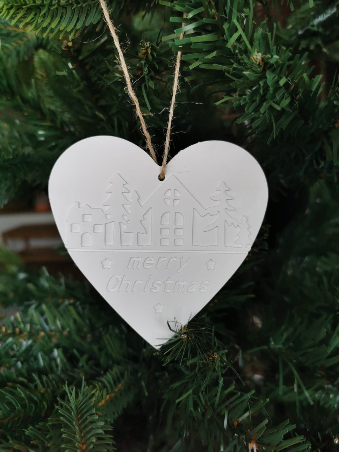 „Weißer Keramik-Weihnachtsanhänger in Herzform mit ‚Merry Christmas‘-Prägung, aufgehängt an einer Juteschnur im Weihnachtsbaum.“