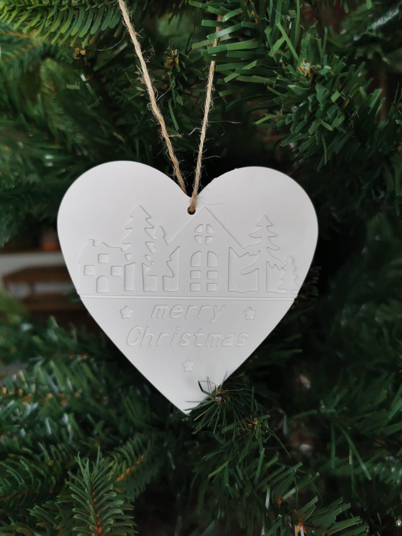 „Weißer Keramik-Weihnachtsanhänger in Herzform mit ‚Merry Christmas‘-Prägung, aufgehängt an einer Juteschnur im Weihnachtsbaum.“