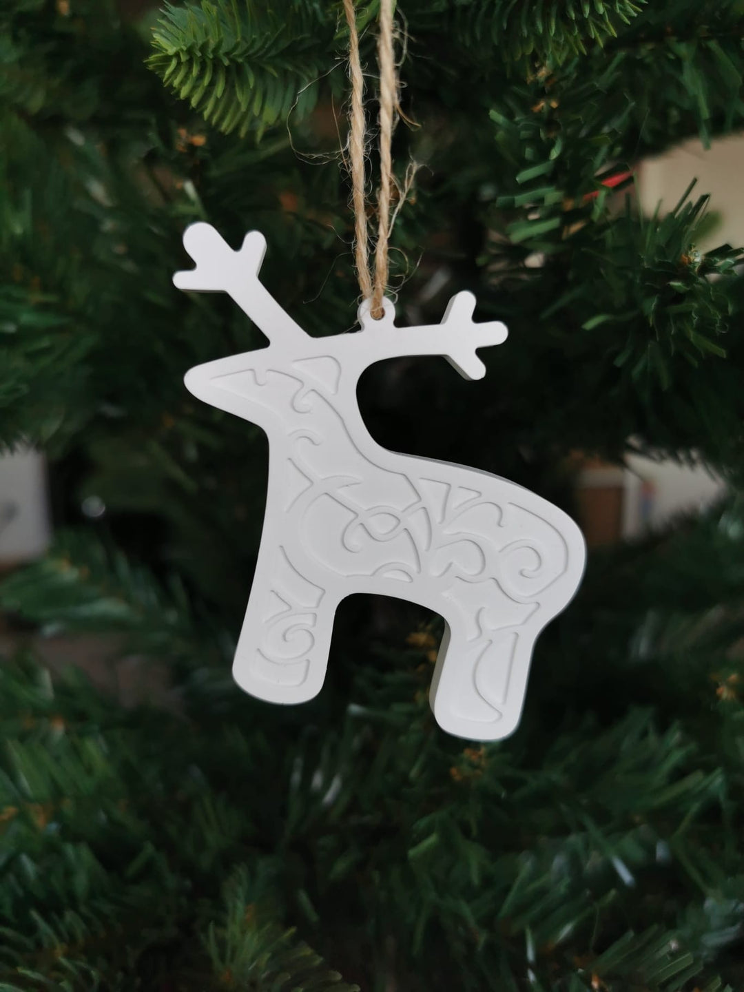 „Weißer Keramik-Baumanhänger in Rentier-Form mit Ornamentmuster, aufgehängt an einer Juteschnur im Weihnachtsbaum.“