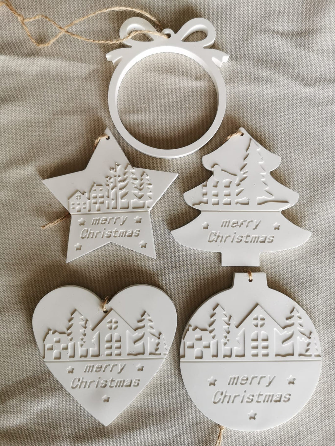 „5-teiliges Set weißer Keramik-Weihnachtsanhänger mit Winter- und Merry-Christmas-Motiven, flach auf Stoff arrangiert.“