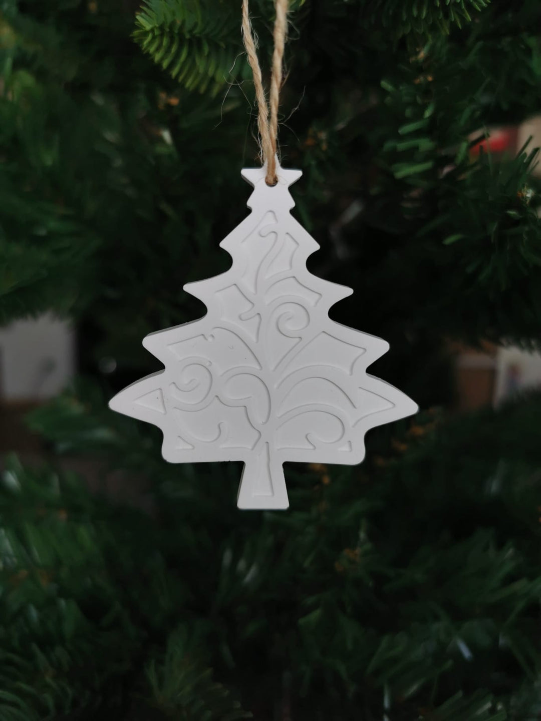 „Weißer Keramik-Baumanhänger in Weihnachtsbaum-Form mit Ornamentmuster, aufgehängt an einer Juteschnur im Tannenbaum.“