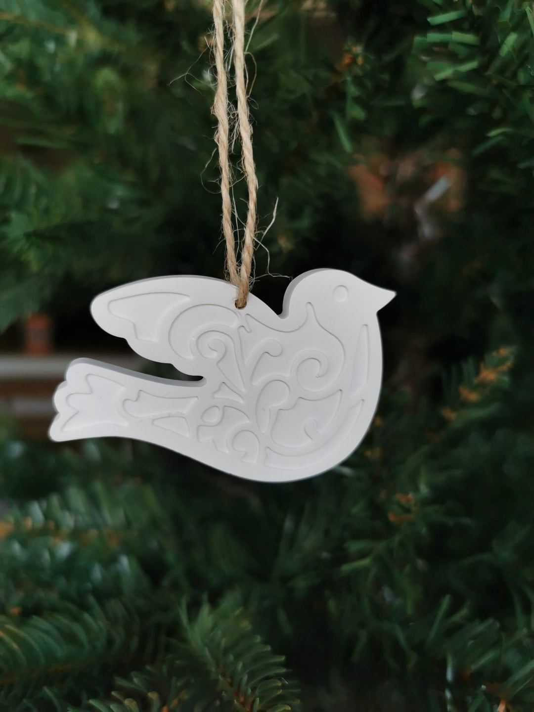 „Weißer Keramik-Baumanhänger in Vogel-Form mit filigranen Ornamenten, aufgehängt an einer Juteschnur in einem Weihnachtsbaum.“