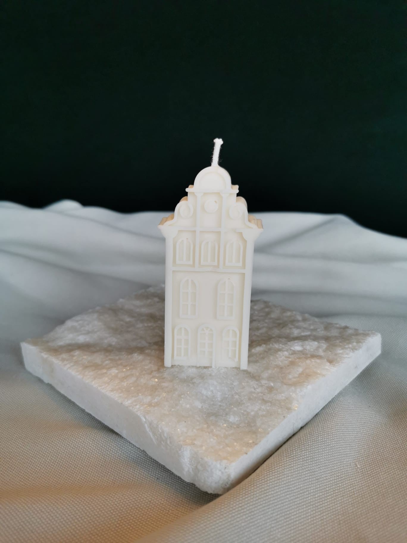„Handgemachte Rapswachskerze in Form eines hohen Stadthauses, stehend auf einer weißen Steinplatte, nachhaltige Deko-Kerze in Cremefarbe.“