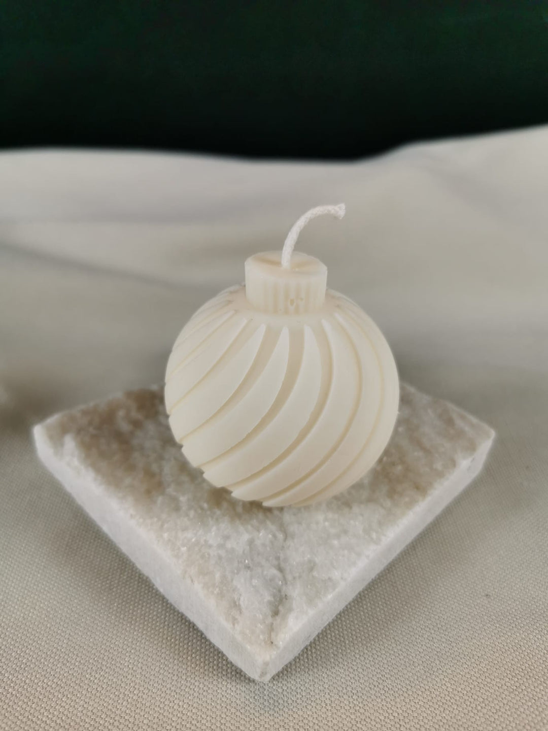 Handgemachte weiße Rapswachskerze in Form einer eleganten Weihnachtskugel mit spiralförmigem Design, fotografiert auf einer hellen Natursteinplatte vor neutralem Hintergrund.