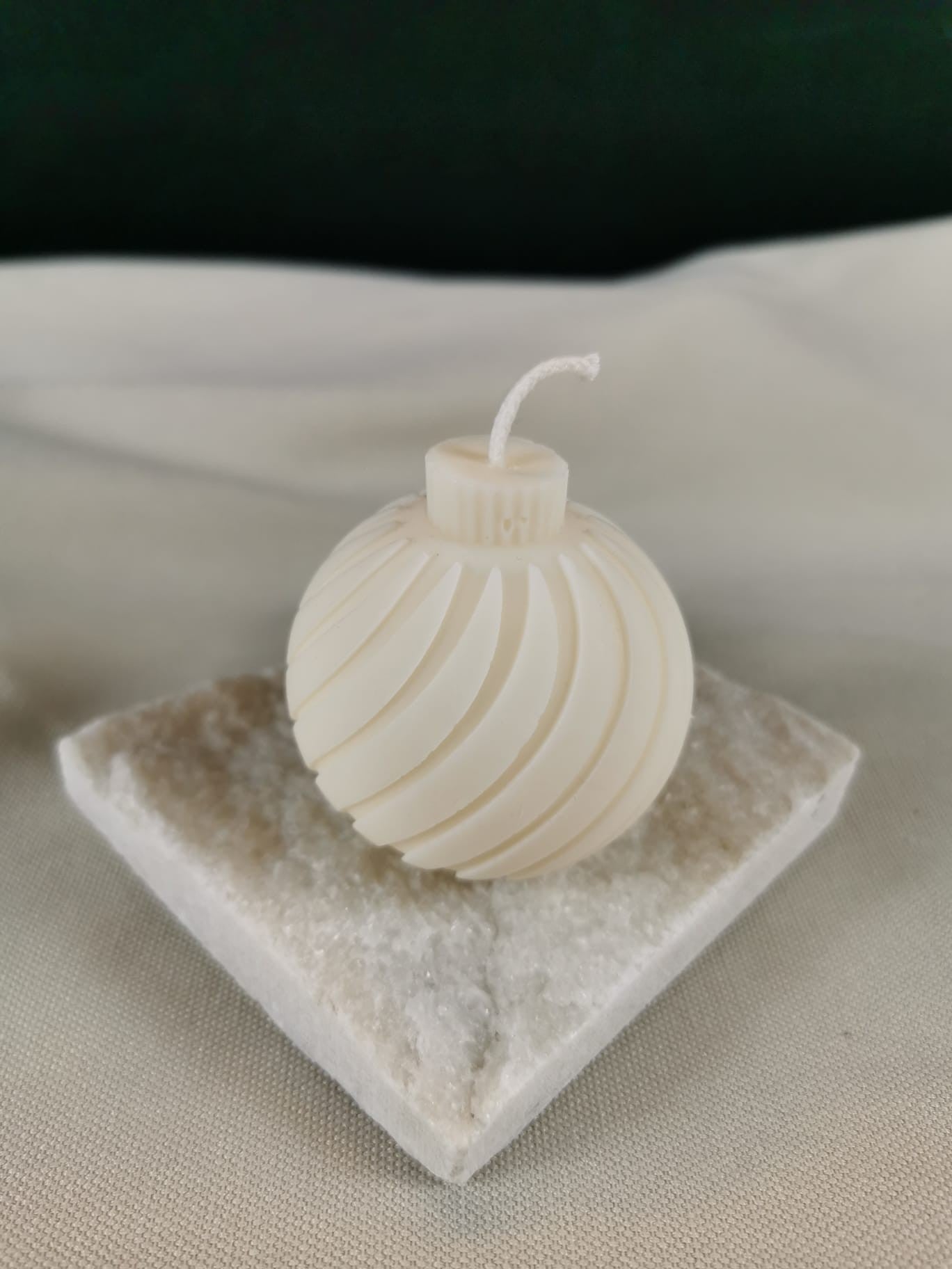 Handgemachte weiße Rapswachskerze in Form einer eleganten Weihnachtskugel mit spiralförmigem Design, fotografiert auf einer hellen Natursteinplatte vor neutralem Hintergrund.