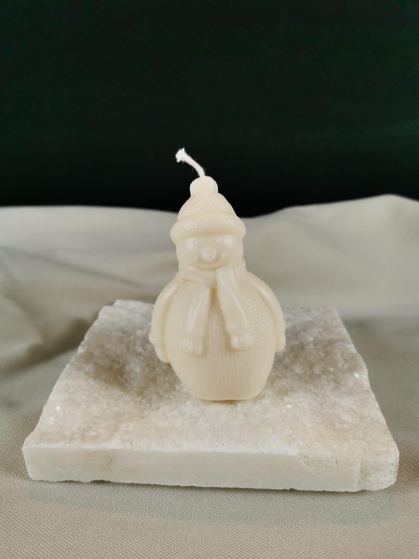 Handgegossene Schneemann-Figurenkerze aus hellem Rapswachs mit Mütze und Schal, dekorativ auf einer glitzernden Steinplatte präsentiert.