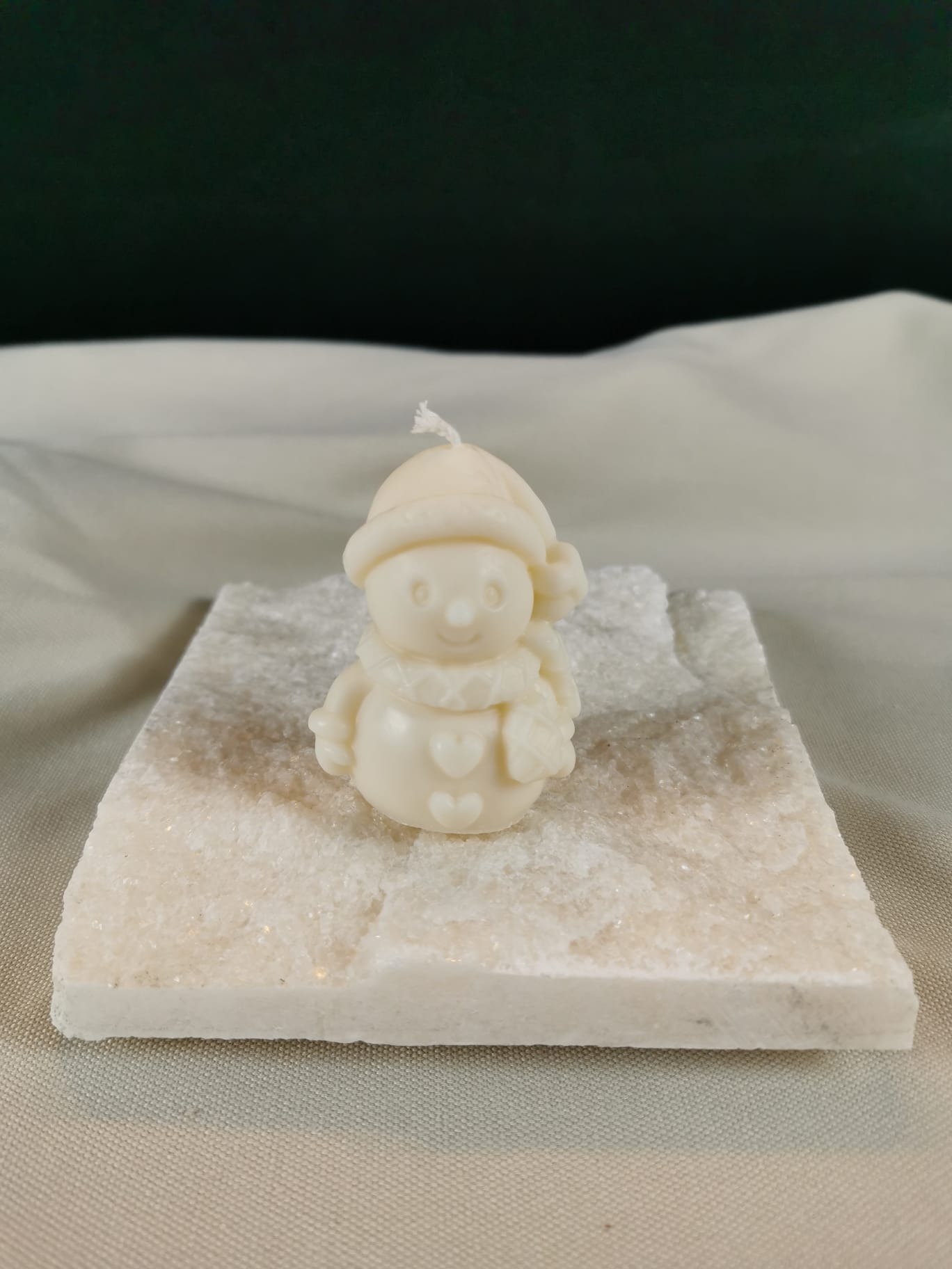 Handgegossene Schneemann-Figurenkerze aus hellem Rapswachs, mit Mütze, Schal und Herzdetails, auf einer glitzernden Steinplatte präsentiert.