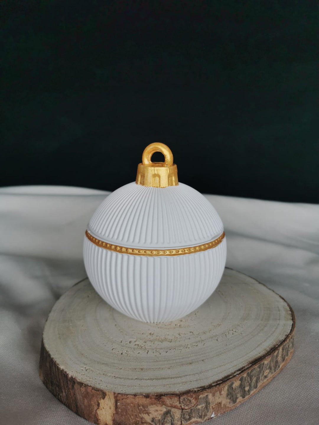 „Edle, weiße Weihnachtskugel aus handgefertigter Keramik mit feinem Rillenmuster und goldenen Details, platziert auf einer Holzscheibe – stilvolle nachhaltige Weihnachtsdekoration.“