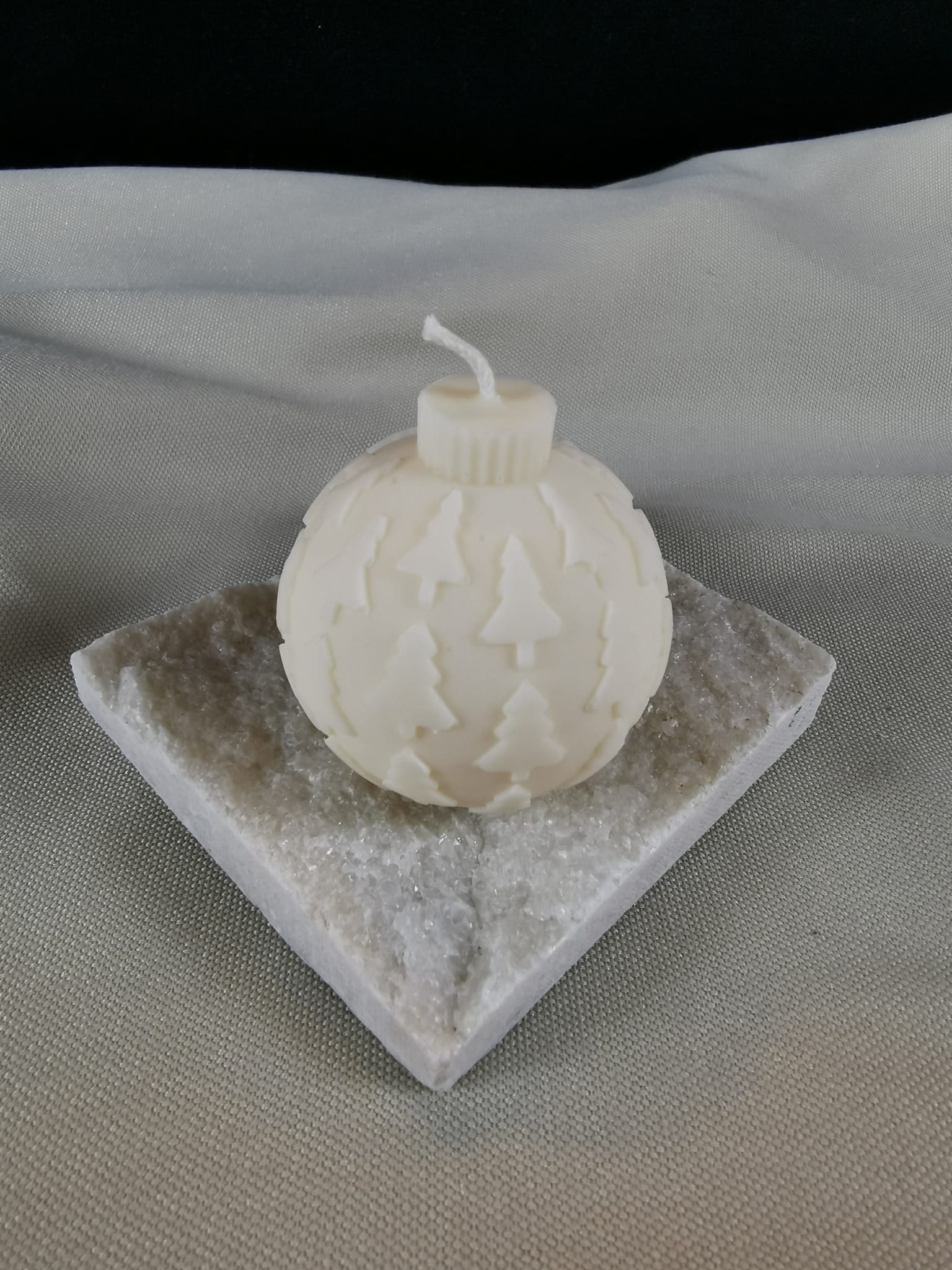 Runde, handgegossene Rapswachs-Kerze in Form einer Weihnachtskugel mit feinem Tannenbaum-Relief, auf einer glitzernden Steinplatte präsentiert.