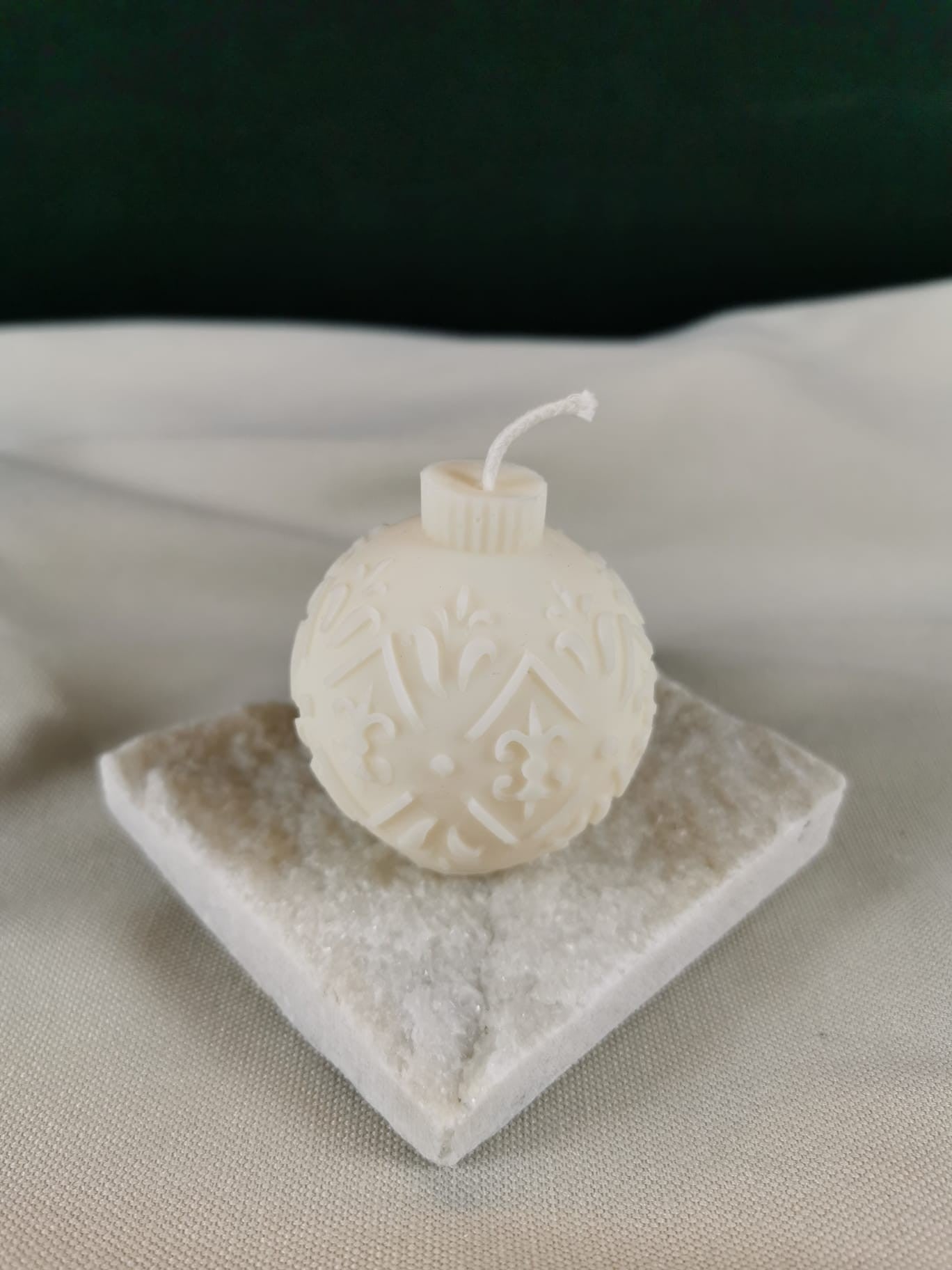 Detailreiche handgegossene Weihnachtskugel-Kerze aus hellem Rapswachs mit ornamentalen Mustern, dekorativ auf Steinplatte präsentiert.