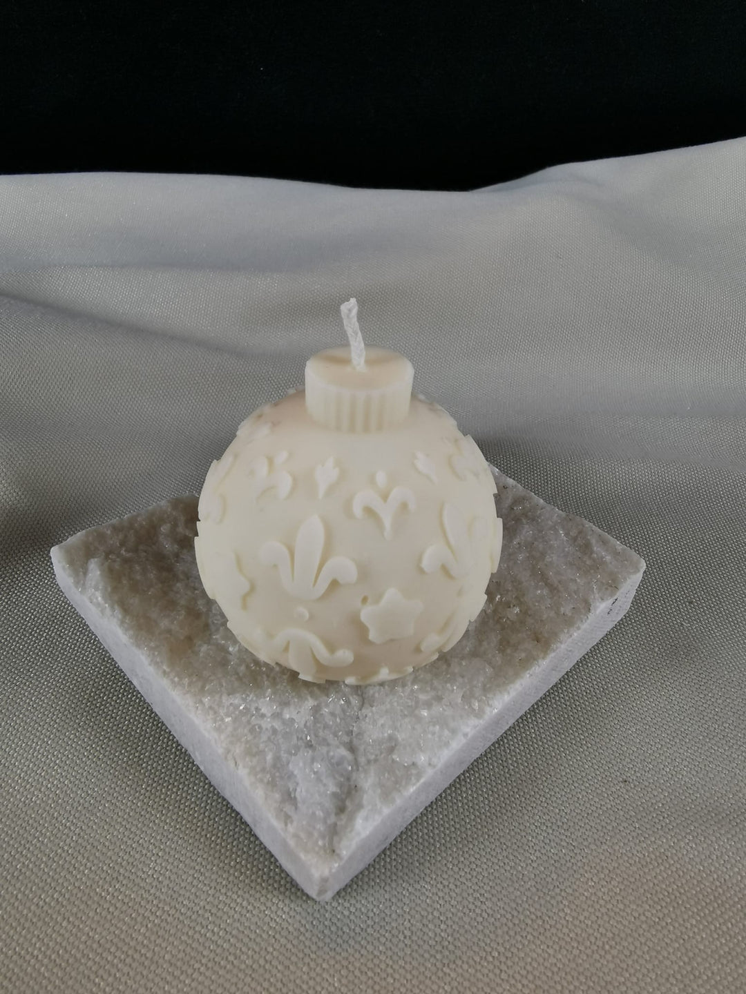 Handgegossene Weihnachtskugel-Kerze aus hellem Rapswachs mit feinen Ornamenten, Sternen und floralen Details, präsentiert auf einer glitzernden Steinplatte.