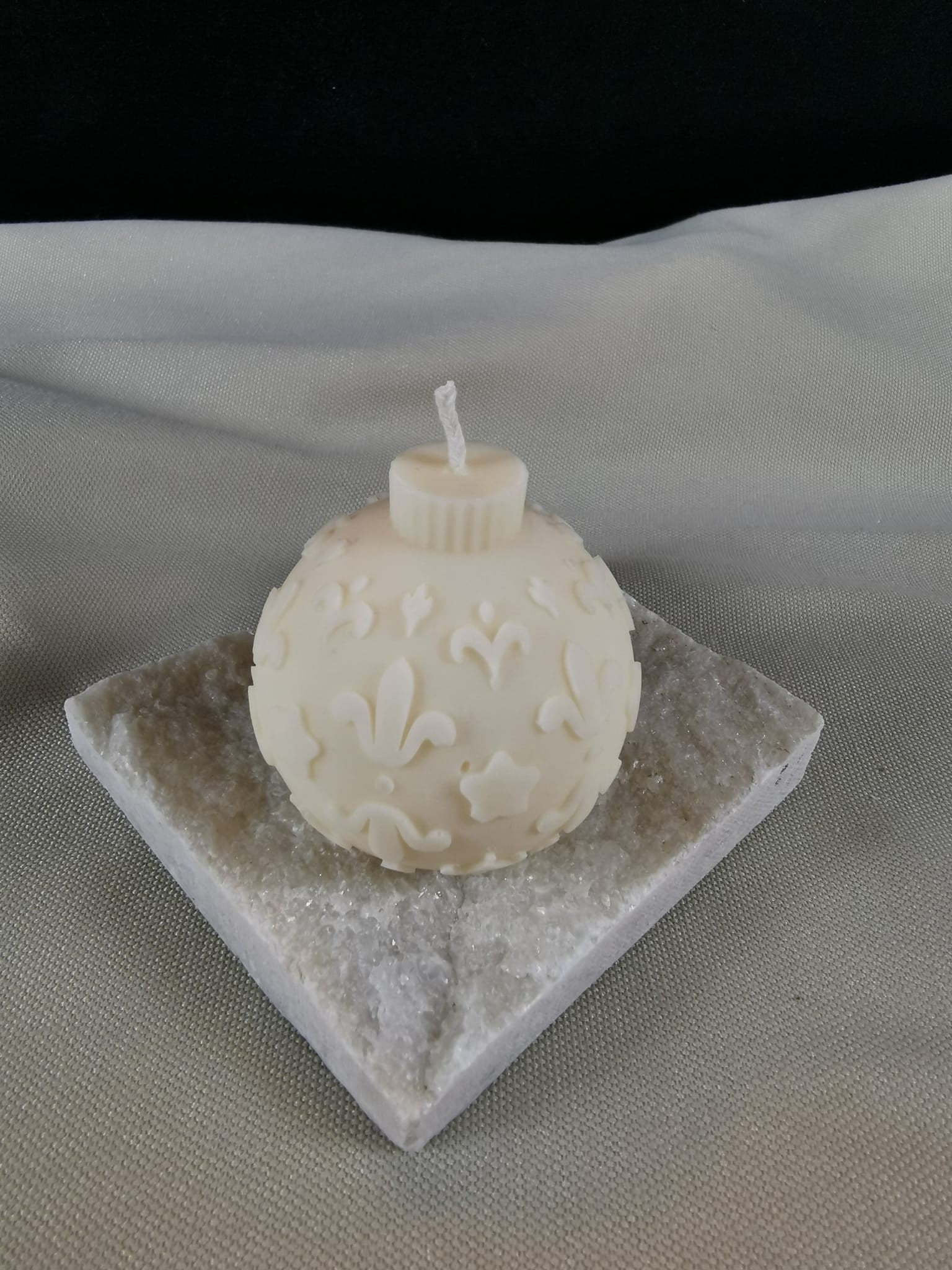 Handgegossene Weihnachtskugel-Kerze aus hellem Rapswachs mit feinen Ornamenten, Sternen und floralen Details, präsentiert auf einer glitzernden Steinplatte.