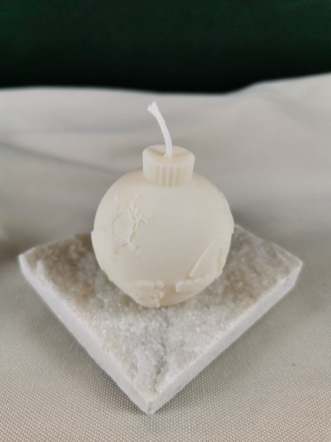 Handgegossene Weihnachtskugel-Kerze aus hellem Rapswachs mit zarter Schneelandschaft und Schneeflockenmotiv, präsentiert auf einer glitzernden Steinplatte.