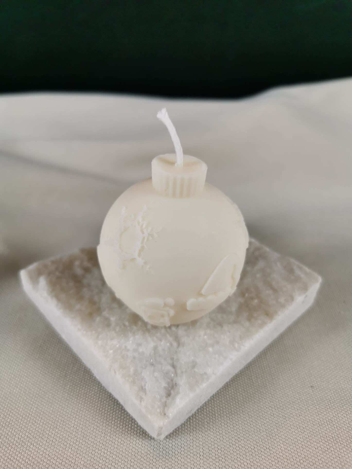 Handgegossene Weihnachtskugel-Kerze aus hellem Rapswachs mit zarter Schneelandschaft und Schneeflockenmotiv, präsentiert auf einer glitzernden Steinplatte.