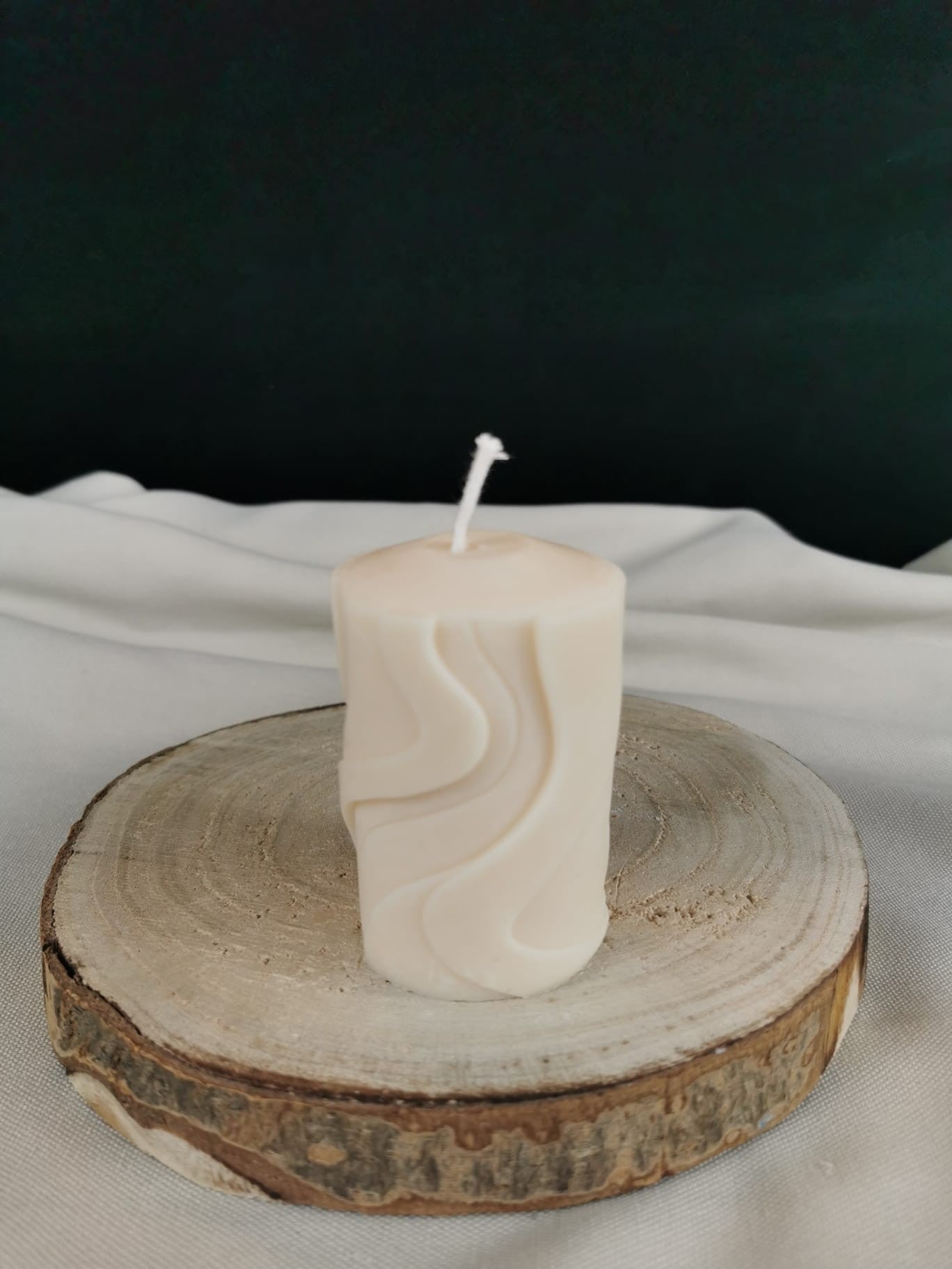 Handgegossene cremefarbene Designkerze in Zylinderform mit elegant geschwungenem Wellenmuster, stehend auf einer Holzscheibe.
